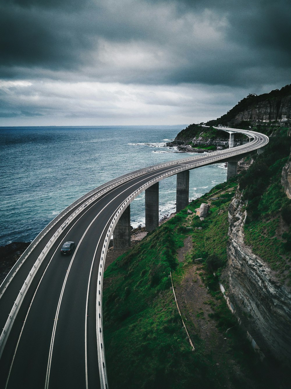 puente de carretera de hormigón gris cerca de la colina y el océano bajo el cielo blanco y azul