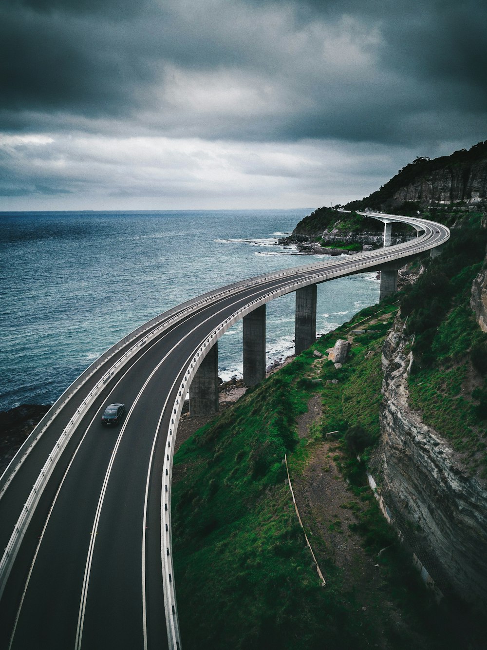 gray concrete road bridge near hill and ocean under white and blue sky