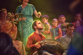 A group of people engaged in a musical performance. The scene includes a person playing a trumpet, another playing what appears to be a percussion instrument, and a person in a teal saree standing with a microphone. Several children are watching in the background, displaying an array of expressions. The setting is warmly lit with colorful lighting effects.