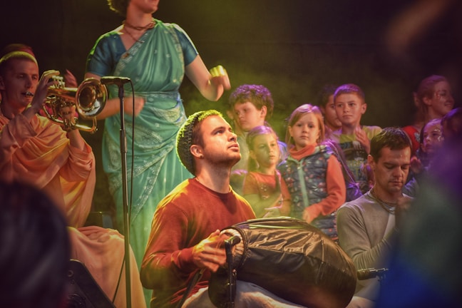 A group of people engaged in a musical performance. The scene includes a person playing a trumpet, another playing what appears to be a percussion instrument, and a person in a teal saree standing with a microphone. Several children are watching in the background, displaying an array of expressions. The setting is warmly lit with colorful lighting effects.