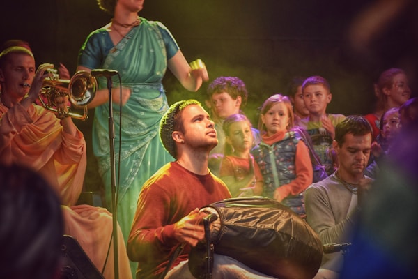 A group of people engaged in a musical performance. The scene includes a person playing a trumpet, another playing what appears to be a percussion instrument, and a person in a teal saree standing with a microphone. Several children are watching in the background, displaying an array of expressions. The setting is warmly lit with colorful lighting effects.