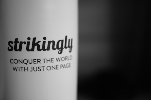 A close-up of black text printed on a white curved surface, with the text reading: 'strikingly' and 'CONQUER THE WORLD WITH JUST ONE PAGE'. The background fades into a dark gradient.