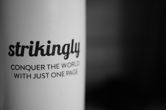 A close-up of black text printed on a white curved surface, with the text reading: 'strikingly' and 'CONQUER THE WORLD WITH JUST ONE PAGE'. The background fades into a dark gradient.