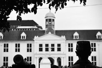 A black-and-white image featuring a historic building with colonial architecture, characterized by white walls and dark roof with decorative windows. In the foreground, a silhouette of a statue or person wearing a hat is visible, alongside what appears to be a cannon. The top is framed by the leaves of a tree.