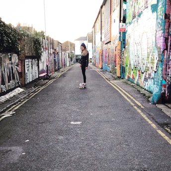 A person is skateboarding down a narrow graffiti-covered alleyway. The walls on both sides are filled with colorful street art, creating a vibrant backdrop. The person is wearing casual clothing and a hooded jacket.