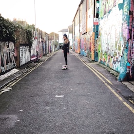 A person is skateboarding down a narrow graffiti-covered alleyway. The walls on both sides are filled with colorful street art, creating a vibrant backdrop. The person is wearing casual clothing and a hooded jacket.