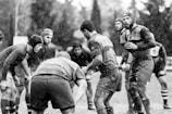 A group of rugby players is gathered closely on a muddy field during a game, as rain falls heavily around them. The players are wearing protective gear such as helmets and jerseys, and their expressions suggest focus and determination.