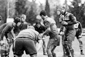 A group of rugby players is gathered closely on a muddy field during a game, as rain falls heavily around them. The players are wearing protective gear such as helmets and jerseys, and their expressions suggest focus and determination.