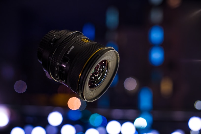 A sleek camera lens focusing sharply against a blurred newsroom background.