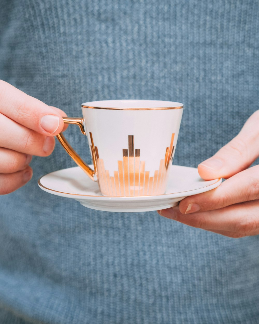 persona que sostiene una taza de té de cerámica blanca y dorada con platillo