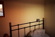 A dimly lit bedroom corner features a metal bed frame with a simple, minimalist design. The bed is unmade, with pillows and sheets casually strewn. A small wall-mounted light fixture is attached to the headboard. A framed window on the left side allows a warm sunset glow to illuminate part of the wall.