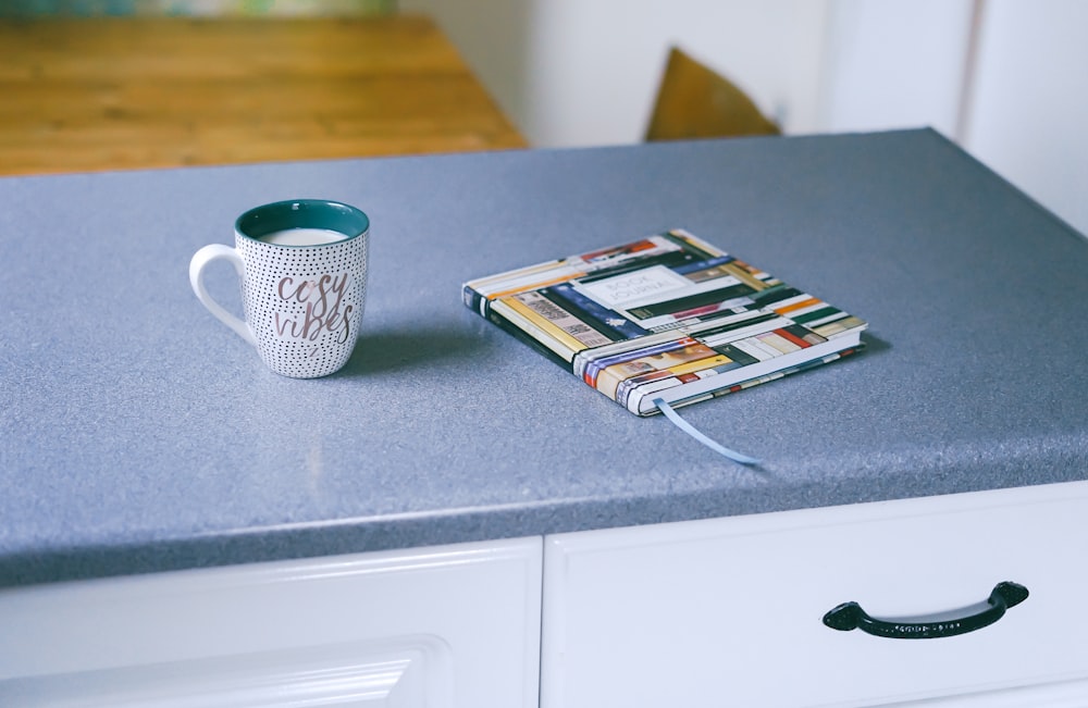 Tasse en céramique grise et blanche à côté d’un livre couvert multicolore