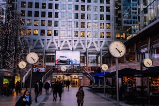 An urban scene at a busy plaza with multiple large clocks displaying time prominently. A modern high-rise building serves as the backdrop, with lit windows indicating activity inside. People are walking through the plaza, some wearing winter coats, suggesting a cold season. A large digital advertisement screen is visible, showing a car. Nearby cafes have outdoor seating under awnings.