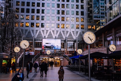 An urban scene at a busy plaza with multiple large clocks displaying time prominently. A modern high-rise building serves as the backdrop, with lit windows indicating activity inside. People are walking through the plaza, some wearing winter coats, suggesting a cold season. A large digital advertisement screen is visible, showing a car. Nearby cafes have outdoor seating under awnings.