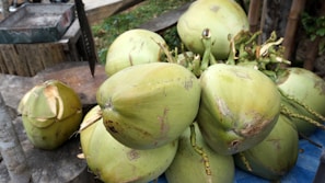 A cluster of fresh green coconuts is placed on a rustic wooden surface with a cutting tool nearby. The coconuts have a natural texture and show some marks and imperfections on their surface. In the background, there are elements of a natural setting like wooden boards and greenery.