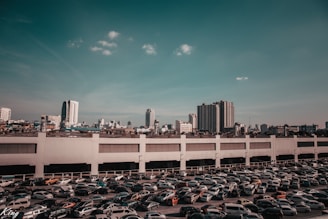 A large parking lot filled with numerous parked cars is situated in front of a tall white building. The background reveals a cityscape with various high-rise buildings under a clear sky with a few scattered clouds.