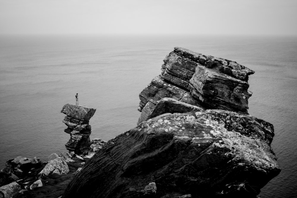 fotografia in scala di grigi di una persona in piedi sulla scogliera vicino all'oceano