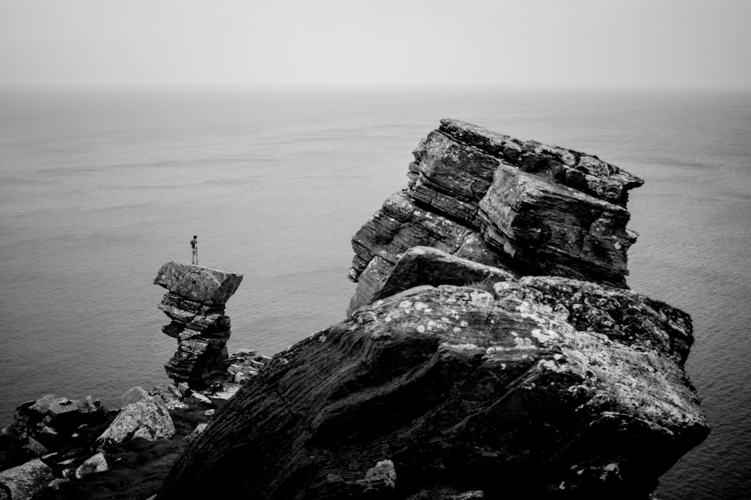 grayscale photography of a person standing on cliff near the ocean