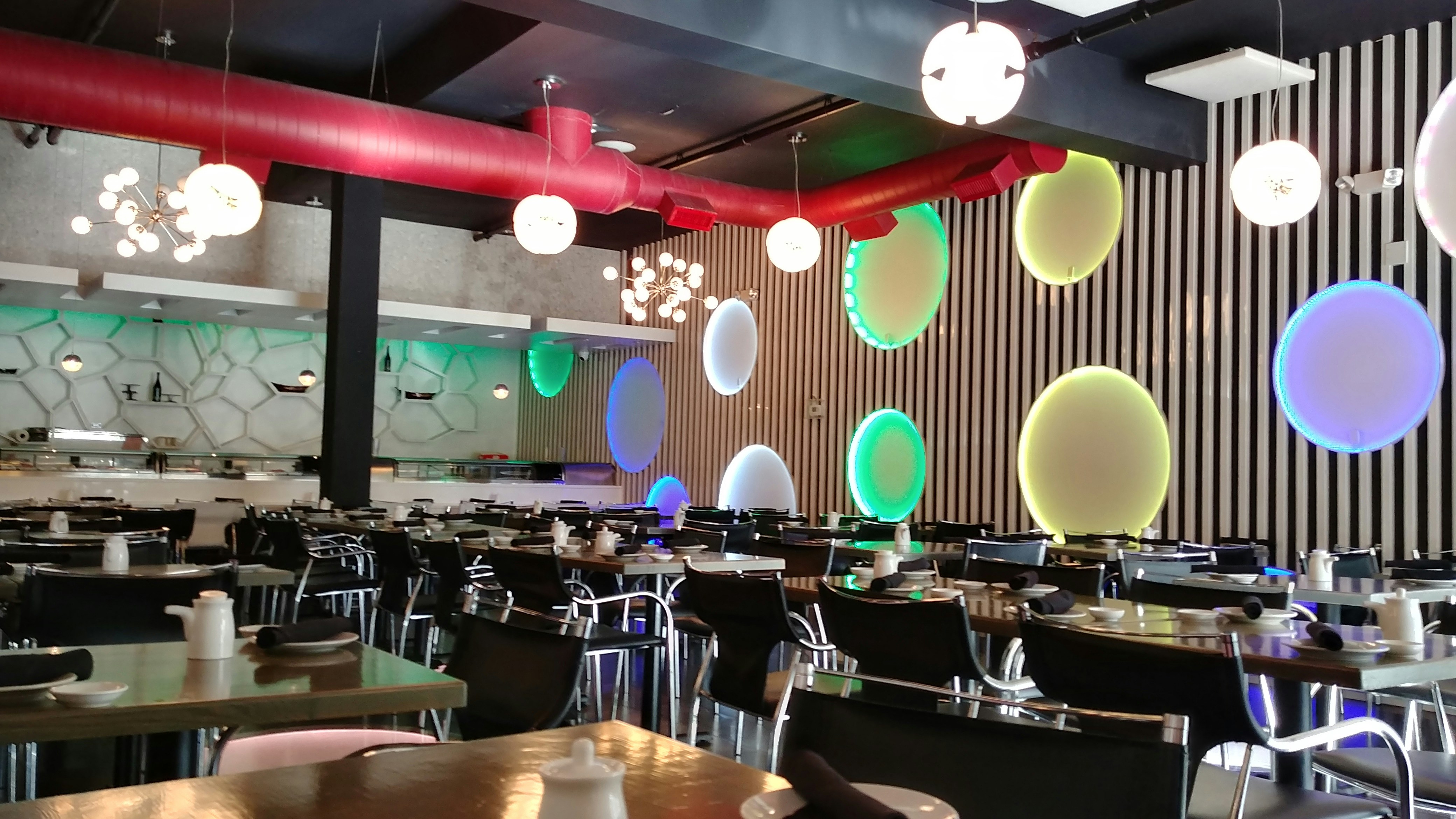 A stylish dining area with modern decor features black tables set with white dishes and utensils. The walls are adorned with circular, multicolored light fixtures and vertical black-and-white stripes. A red ventilation pipe runs across the ceiling, complementing the contemporary light fixtures hanging from above.
