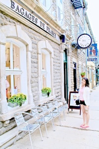 A stone building with signage 'Bagages de France' features a row of metallic chairs along the sidewalk. Windows with flower boxes are integrated into the structure. A woman in casual attire, holding a cup, stands facing the camera next to a framed poster. The area has a sunny ambiance, and there are additional signs visible in the background indicating a hotel.