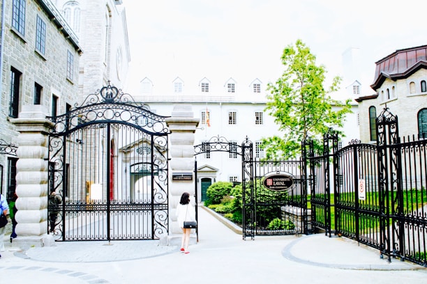 An ornate wrought iron gate stands open, leading into a courtyard flanked by historic stone buildings with multiple windows. A person walks towards the gate, dressed casually, as a tree with lush green leaves stands nearby. The atmosphere suggests a blend of academia and history, with well-maintained architecture and landscaping.