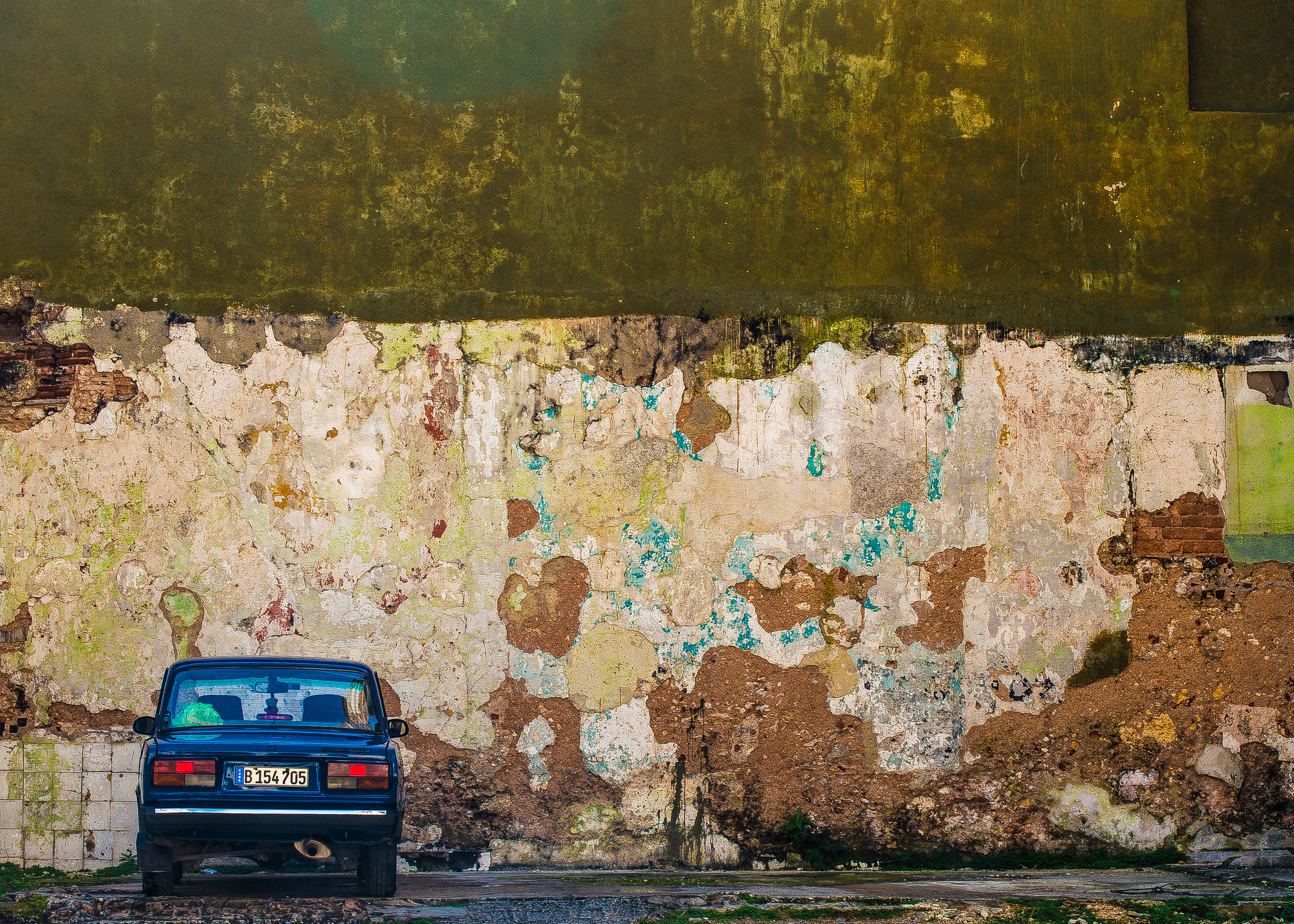 A vintage blue car is parked against a heavily weathered, peeling painted wall.