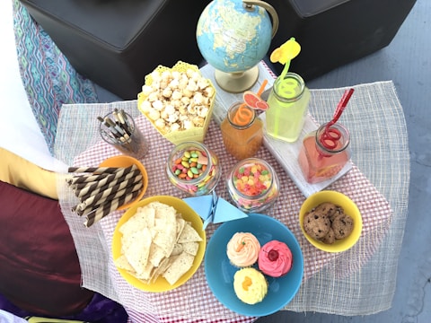 An assortment of snacks and drinks is arranged on a table with a checkered cloth. There's a globe, popcorn in a decorative container, glasses with colorful drinks and fun straws, jars of assorted candies, striped wafers, a blue plate with cupcakes, and a yellow bowl with cookies and tortilla chips.