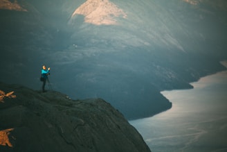 A person stands on a rocky cliff taking photographs with a tripod, overlooking a vast, tranquil landscape that includes a body of water bordered by rugged mountains. The light creates a serene atmosphere.