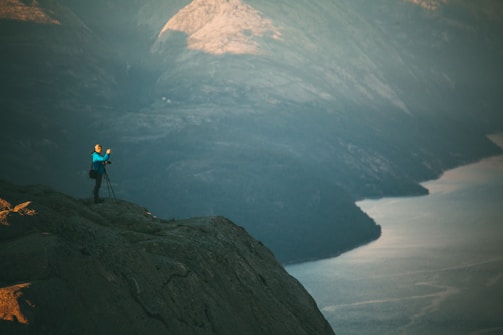 A person stands on a rocky cliff taking photographs with a tripod, overlooking a vast, tranquil landscape that includes a body of water bordered by rugged mountains. The light creates a serene atmosphere.
