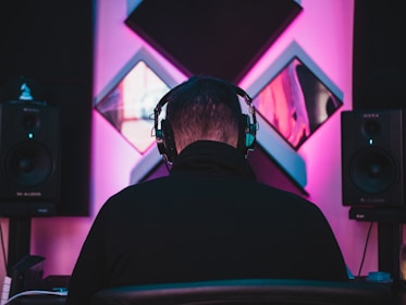 A person wearing headphones is seated facing a wall adorned with acoustic panels. Two large speakers are positioned on either side, emitting soft ambient lighting in shades of pink and blue, creating a moody studio atmosphere.