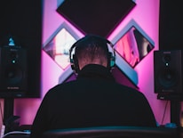 A person wearing headphones is seated facing a wall adorned with acoustic panels. Two large speakers are positioned on either side, emitting soft ambient lighting in shades of pink and blue, creating a moody studio atmosphere.