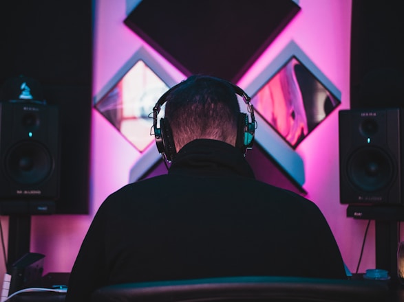 A person wearing headphones is seated facing a wall adorned with acoustic panels. Two large speakers are positioned on either side, emitting soft ambient lighting in shades of pink and blue, creating a moody studio atmosphere.