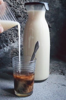 A glass is being filled with milk pouring from a small container, creating a swirling pattern with the dark coffee inside. Next to it, a tall carafe filled with milk stands on a textured concrete surface, with a spoon inside the glass.