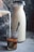 A glass is being filled with milk pouring from a small container, creating a swirling pattern with the dark coffee inside. Next to it, a tall carafe filled with milk stands on a textured concrete surface, with a spoon inside the glass.