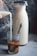 A glass is being filled with milk pouring from a small container, creating a swirling pattern with the dark coffee inside. Next to it, a tall carafe filled with milk stands on a textured concrete surface, with a spoon inside the glass.