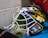 A colorful hockey goalie mask with a white cage and vibrant blue, yellow, and silver design. It sits on a blue surface, and there is a roll of tape inside. Part of a red sleeve is visible in the upper right corner.