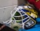 A colorful hockey goalie mask with a white cage and vibrant blue, yellow, and silver design. It sits on a blue surface, and there is a roll of tape inside. Part of a red sleeve is visible in the upper right corner.