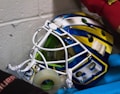 A colorful hockey goalie mask with a white cage and vibrant blue, yellow, and silver design. It sits on a blue surface, and there is a roll of tape inside. Part of a red sleeve is visible in the upper right corner.