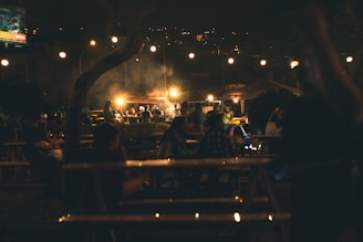 Guests laughing and sharing food around a long picnic table under string lights at sunset.