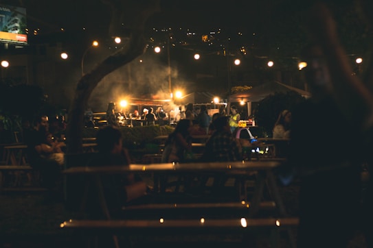 A nighttime outdoor gathering featuring people seated at picnic tables, illuminated by hanging string lights. A smoky atmosphere surrounds the area, likely from food preparation, as people interact in a lively social setting.
