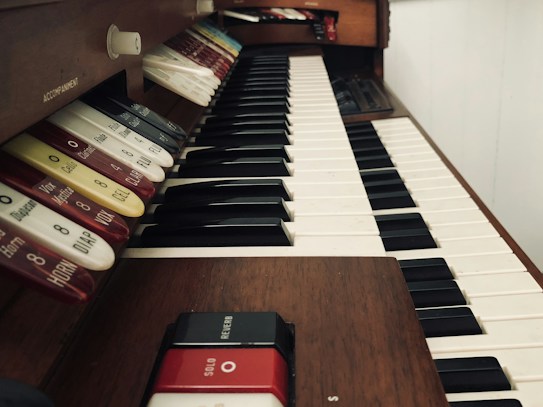 An organ keyboard with multiple keys and stops, showcasing wooden paneling and a range of labeled switches for different musical effects. The perspective is angled, focusing on the arrangement of the keys and the adjacent control mechanisms.