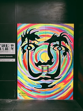 A vibrant mural art piece featuring a stylized face with large eyes and abstract features. The face is outlined in black against a swirling, colorful background of rainbow-like patterns. The artwork gives a psychedelic visual effect with its bold lines and curves.