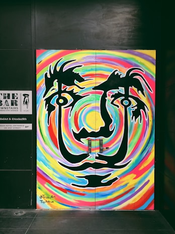 A vibrant mural art piece featuring a stylized face with large eyes and abstract features. The face is outlined in black against a swirling, colorful background of rainbow-like patterns. The artwork gives a psychedelic visual effect with its bold lines and curves.