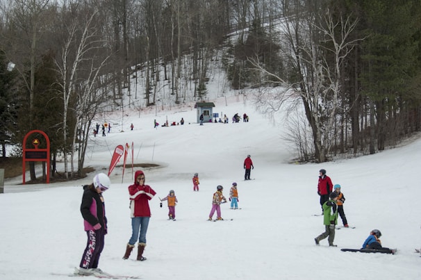 A snowy landscape with several people skiing and playing in the snow. Children and adults in colorful winter clothing are scattered across the slope, indicating a day of leisure and fun in a ski resort. Bare trees surround the area, with a small building visible in the background. Red banners and ski equipment are present, adding vibrant touches to the serene winter scene.