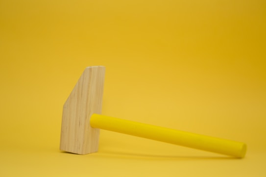 A wooden mallet with a yellow handle rests against a seamless yellow background, creating a minimalist and monochromatic aesthetic.