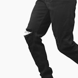 A close-up of a person wearing black ripped jeans, with visible frayed edges around the tears on both knees. The image focuses on the texture and style of the jeans against a plain white background.