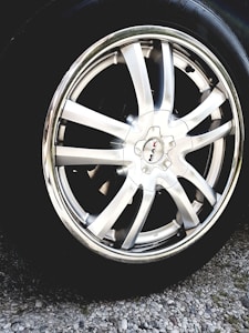 A close-up view of a shiny silver alloy wheel rim with multiple broad spokes. The wheel is mounted on a tire and is surrounded by gravel on the ground. The center cap of the wheel has a logo with the text 'MAK'. The metallic sheen reflects light, giving a polished appearance.