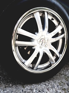A close-up view of a shiny silver alloy wheel rim with multiple broad spokes. The wheel is mounted on a tire and is surrounded by gravel on the ground. The center cap of the wheel has a logo with the text 'MAK'. The metallic sheen reflects light, giving a polished appearance.