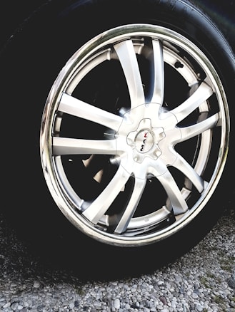 A close-up view of a shiny silver alloy wheel rim with multiple broad spokes. The wheel is mounted on a tire and is surrounded by gravel on the ground. The center cap of the wheel has a logo with the text 'MAK'. The metallic sheen reflects light, giving a polished appearance.