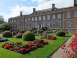 A large brick manor house stands in the background with numerous windows and chimneys. In front of the building, there is an intricately landscaped garden featuring vibrant green lawns, neatly trimmed spherical bushes, and colorful flowerbeds predominantly with red flowers. A stone pathway runs through the garden, enhancing its symmetry and aesthetic appeal.